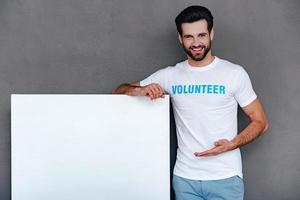 We can help to community Confident young man in volunteer t-shirt pointing on white boardand looking at camera with smile while standing against grey background photo