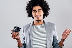 quiero una foto de un joven africano apuesto que sostiene una cámara de estilo retro y mira la cámara con una sonrisa mientras se enfrenta a un fondo gris