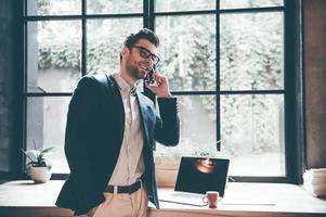 Always in touch with colleagues. Confident young man talking on the mobile phone and smiling while standing in front of the big window in office or cafe photo