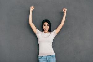 ella es una mujer joven feliz ganadora en ropa casual manteniendo los brazos levantados y mirando a la cámara mientras está de pie contra el fondo gris foto