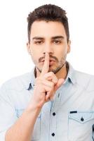 Keep my secret Serious young man in shirt holding finger on lips and looking at camera while standing against white background photo