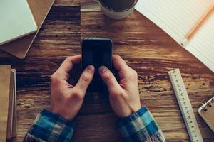 Texting business message. Close-up top view of male hands holding mobile phone over wooden desk with different chancellery stuff laying on it photo