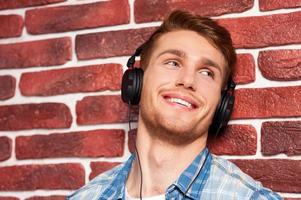 Music is my life. Handsome young man listening to the MP3 Player and looking away while standing against brick wall photo