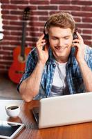 disfrutando de la musica vista superior de un joven apuesto con auriculares que trabaja en una laptop y sonríe mientras la guitarra acústica se pone en el fondo foto