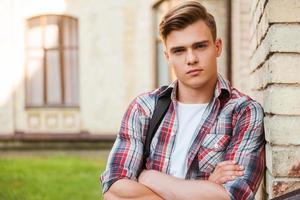 Confident college student. Confident young man carrying backpack on one shoulder and keeping arms crossed while leaning at the brick wall photo