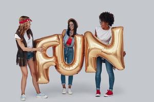 Full length of three attractive young women carrying balloons and smiling while standing against grey background photo