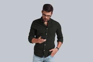 Quick message to client.  Handsome young man in smart casual wear holding smart phone and looking at it while standing against grey background photo