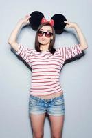 Playful mouse.  Playful young woman in glasses holding hands on fake ears and making a face while standing against grey background photo