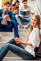 Relaxing before the next class. Low angle view of happy young woman holding digital tablet and looking at camera while people sitting in the background photo
