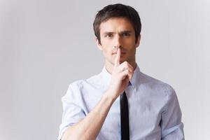 Keep a secret Confident young man in white shirt looking at camera and holding finger on lips while standing against grey background photo