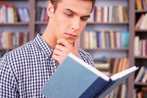 leyendo su libro favorito. joven concentrado leyendo un libro y sosteniendo la mano en la barbilla mientras está de pie contra la estantería foto