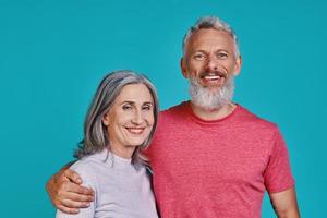 Happy senior couple looking at camera and smiling while standing together against blue background photo