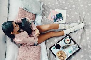 Top view of beautiful young woman in pajamas embracing domestic cat while resting in bed at home photo