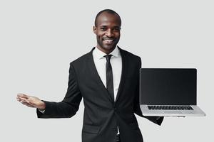 joven africano moderno con ropa formal que señala el espacio de la copia en la computadora portátil y sonríe mientras está de pie contra el fondo gris foto