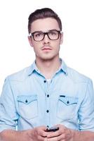 Smart and confident. Handsome young man in shirt looking at camera and holding mobile phone  while standing against white background photo