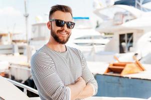 Confident yachtsman. Smiling young man keeping arms crossed and looking at camera while standing on the board of yacht photo