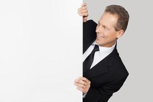 Looking at your text. Cheerful mature man in formalwear looking out of copy space and looking at it while standing against grey background photo