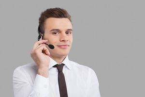 Man in headset. Cheerful young man in formalwear adjusting his headset and smiling at camera while standing against grey background photo