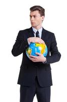 World in his hands. Confident young man in formalwear holding globe and looking away while standing isolated on white photo