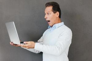 What is that Surprised mature man looking at laptop and keeping mouth open while standing against grey background photo