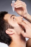 Taking care of his eyes. Side view of young man applying eye drops while standing against grey background photo