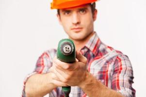 Freeze Handsome young handyman in hardhat stretching out drill and looking at camera while standing against grey background photo