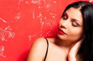 Beauty on red. Portrait of beautiful young woman keeping eyes closed and touching her neck with hand while standing against red background photo