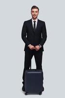 Business travel. Full length of good looking young man with suitcase smiling and looking at camera while standing against grey background photo