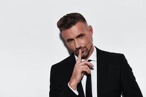 Can you keep a secret Handsome young man in full suit keeping index finger by his lips and smiling while standing against white background photo