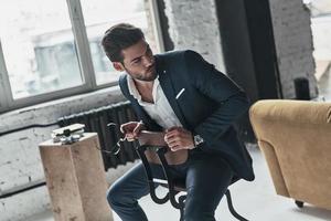Confident young man. Handsome young man in full suit looking away while sitting on the stool photo