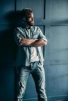 Irresistible man. Confident young bearded man in blue jeans shirt looking away and keeping arms crossed while standing against wall photo