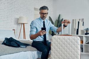 Handsome mature man in shirt and tie leaning at the suitcase while sitting on bed at home photo