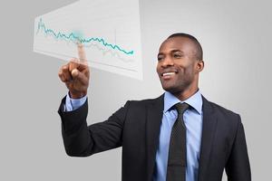 Innovative business. Cheerful young African man in formalwear pointing diagram on the transparent wipe board while standing against grey background photo