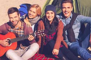 Spending great time with friends. Top view of four young happy people sitting near the tent together while young handsome man playing guitar and smiling photo