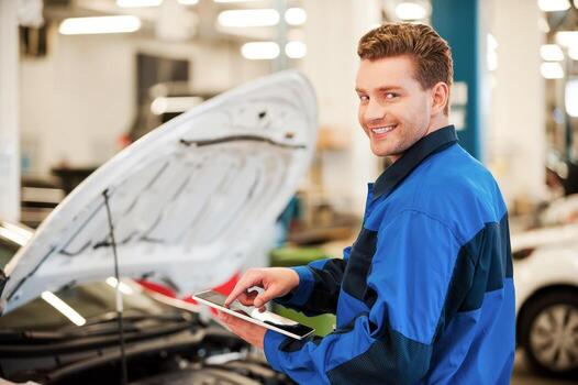 Years of mechanical experience. Confident young man working on digital tablet and smiling while standing in workshop with car in the background photo