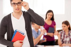 Confident and smart student. Confident male student holding books and adjusting his glasses while other students sitting on background photo