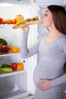 Hungry pregnant woman. Beautiful young pregnant woman standing near the open fridge and eating sandwich photo