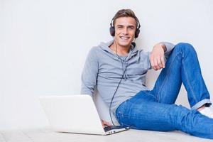 Carefree and relaxed. Happy young man in headphones listening to the music and looking at you while sitting on the floor with laptop near him photo