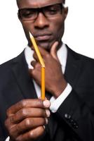 I need a new one. Thoughtful young African man in formalwear holding pencil and looking at it while holding hand on chin and standing isolated on white background photo