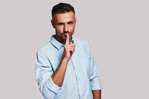 Shhh... Thoughtful young man keeping index finger by his lips and looking at camera while standing against grey background photo