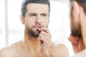 Painful procedure. Concentrated young man tweezing hair from nose and looking at himself while standing in front of the mirror photo