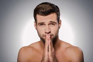 Begging for... Portrait of frustrated young shirtless man looking at camera and keeping and clasped near face while standing against grey background photo