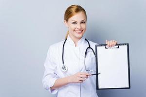 Everything is ok, here you can read your diagnose. Confident female doctor in white uniform outstretching clipboard and looking at camera while standing against grey background photo