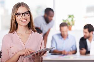 Confident in her team. Confident young woman in glasses working on digital tablet and smiling while three people working on background photo