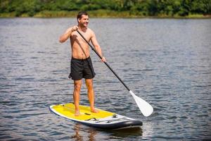 Relaxing on paddleboard. Handsome young man surfing on his paddleboard and smiling photo