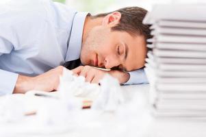 agotado y con exceso de trabajo. un joven apuesto con camisa y corbata durmiendo sentado en la mesa con un montón de papeleo encima foto