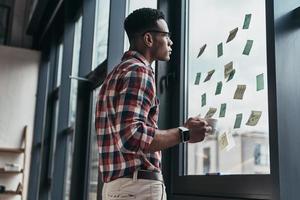 lluvia de ideas. joven serio que aplica notas adhesivas para construir una estrategia de negocios mientras está de pie cerca de la ventana foto