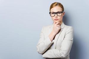 Thoughtful businesswoman. Confident young businesswoman holding hand on chin and looking at camera while standing against grey background photo