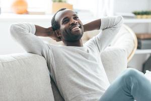 descansando en casa. un apuesto joven africano cogido de la mano detrás de la cabeza y sonriendo mientras se sienta en el sofá de casa foto