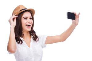 Me encanta la selfie alegre mujer joven con sombrero funky haciendo selfie con su teléfono inteligente y sonriendo mientras está de pie contra el fondo blanco. foto